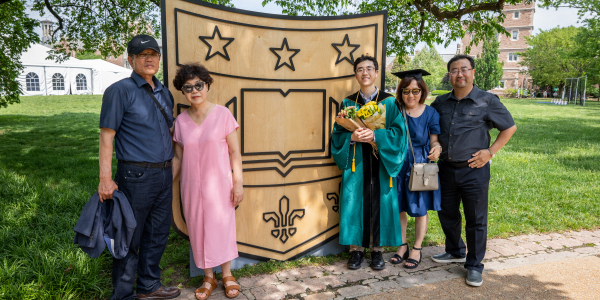 Washington University in St. Louis graduating student and family members pose in front of a prominent WashU shield emblem, symbolizing their pride and celebration of academic accomplishment.