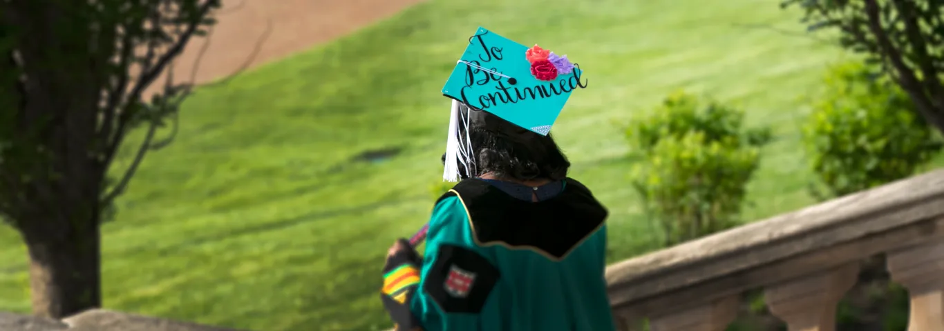 Washington University in St. Louis graduating student in academic attire, including a cap adorned with flowers and the words 'to be continued,' descends outdoor campus stairs, symbolizing their transition to future opportunities and achievements.